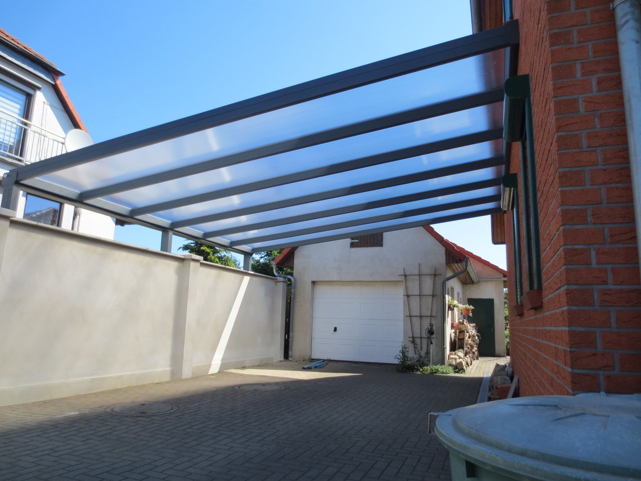 Ein Carport mit transparentem Dach und grauem Metallrahmen neben einem Backsteinhaus und einer Garage.