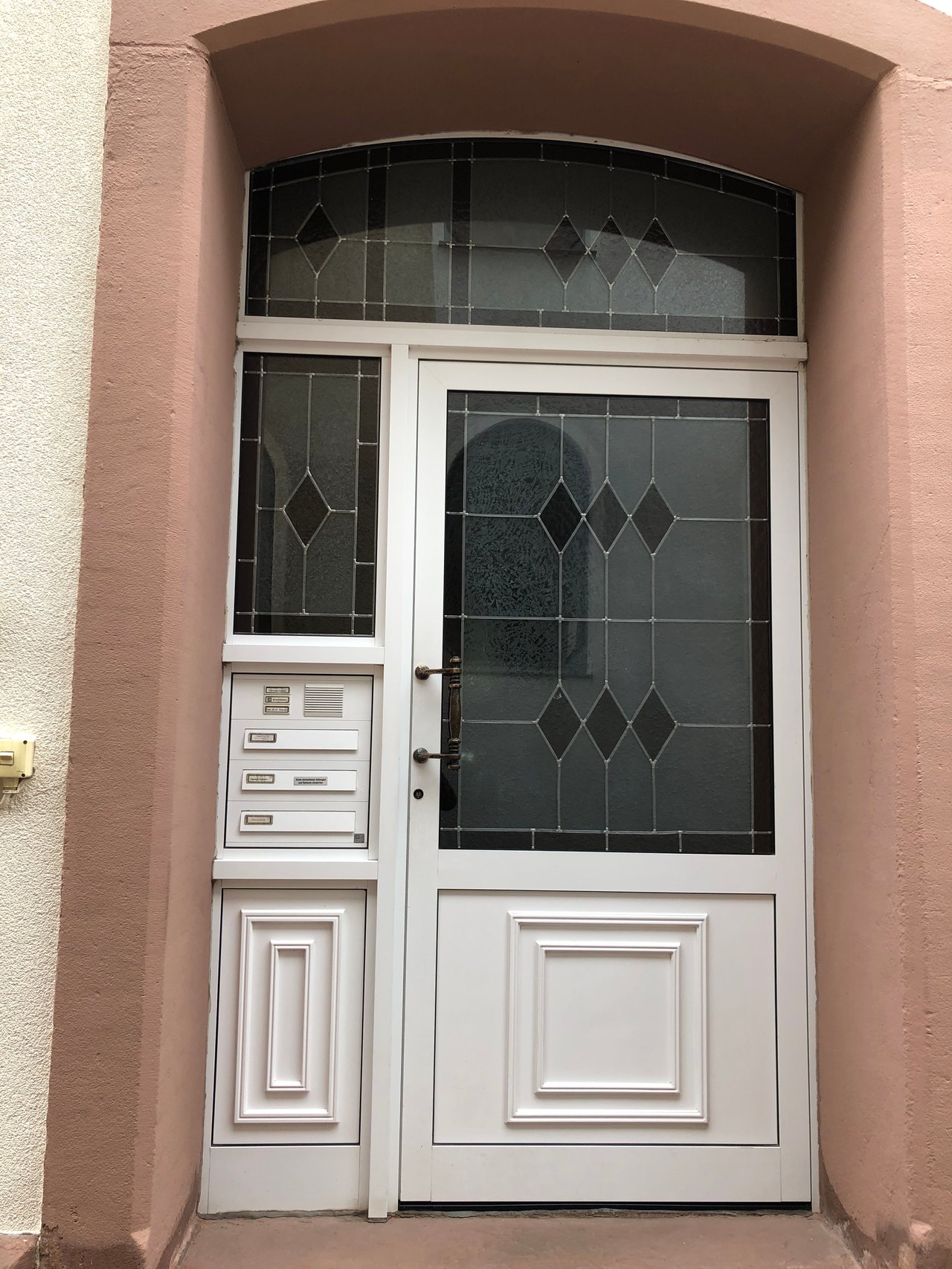 Weiße Haustür mit Glaseinsätzen, einem Seitenlicht und drei Briefkästen in einer rosa Fassade.