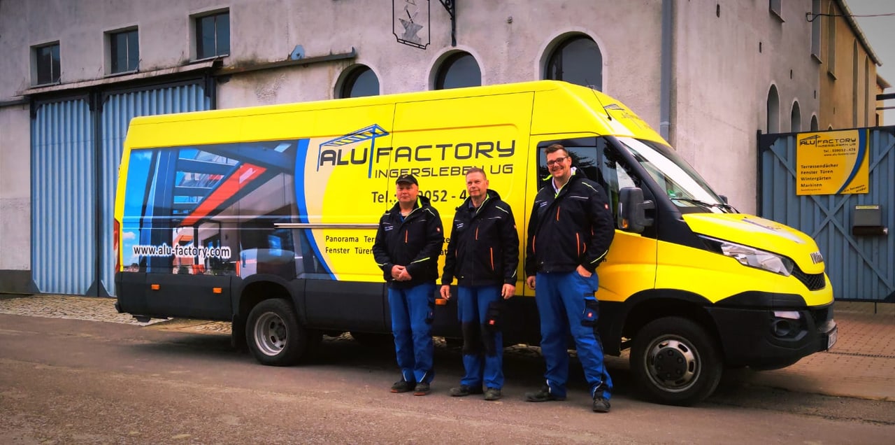 Drei Männer stehen vor einem gelben Lieferwagen mit dem "ALUFACTORY" Logo.